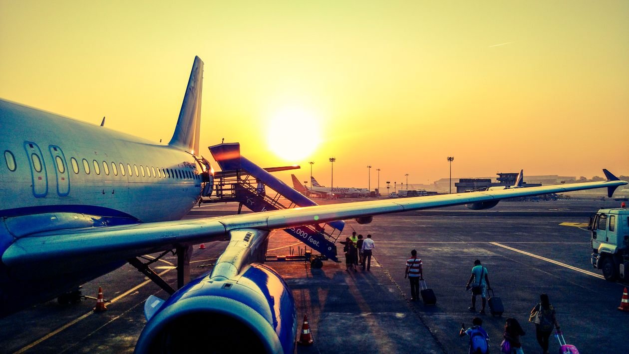 Passengers walking across the airport apron toward an aircraft at sunset, with the plane's wing and fuselage in the foreground