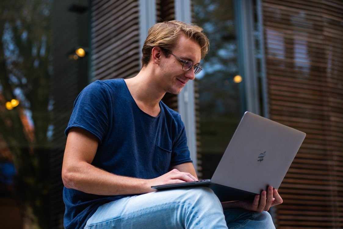 A young adult English learner in glasses sitting outdoors, reading and studying on a laptop