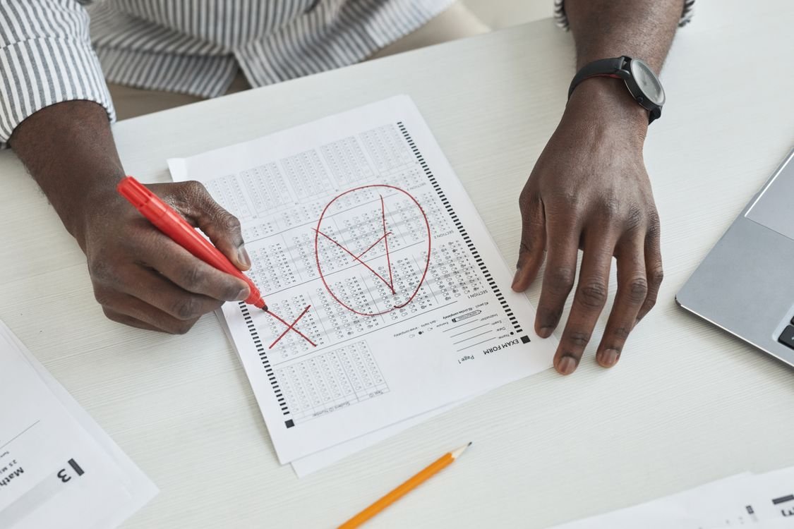 A hand marking a multiple-choice exam answer sheet with a red marker, drawing a large tick and a cross on the EXAM FORM, with a laptop beside it on a desk