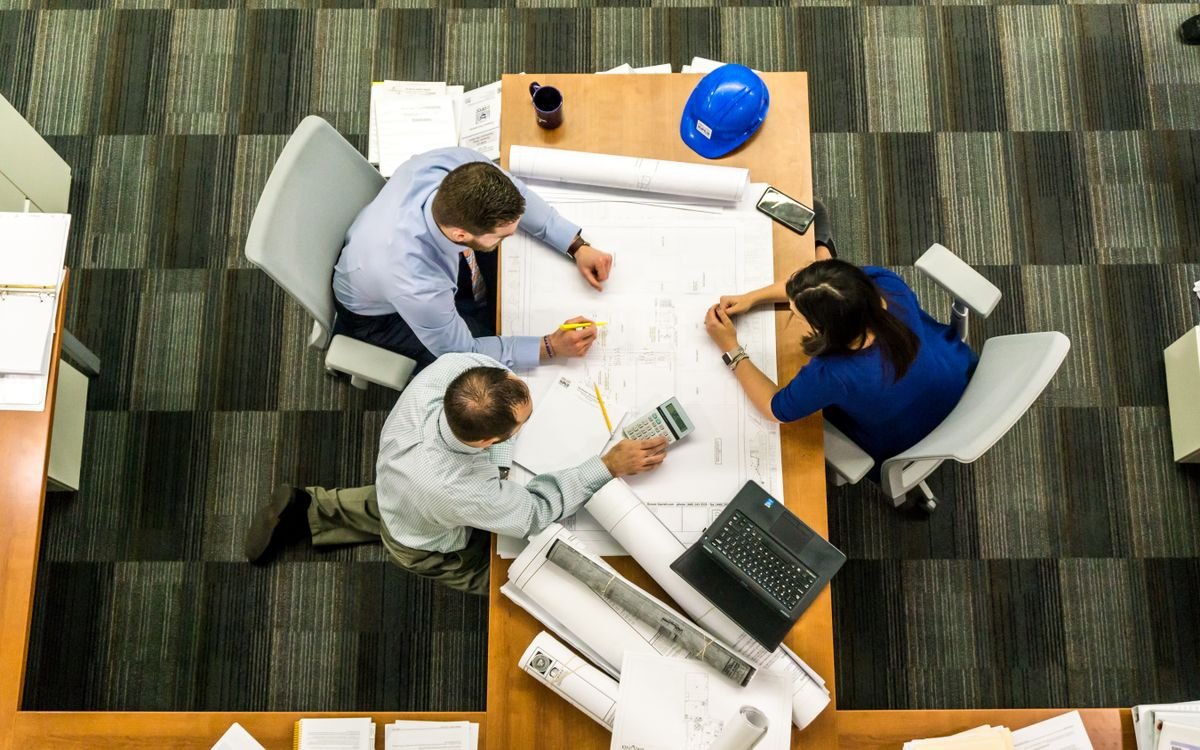 Overhead view of three architects and designers at a desk reviewing building blueprints, with a hard hat, laptop, calculator and rolled technical drawings