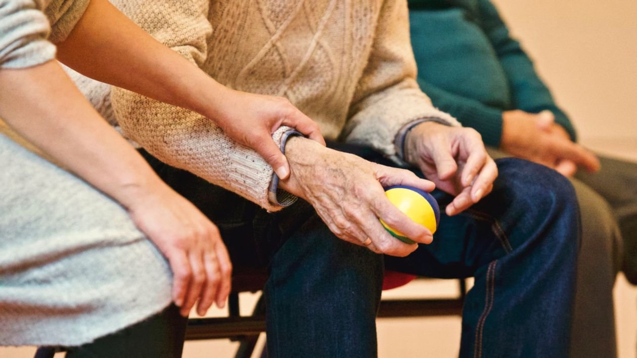 A care worker gently supporting an elderly person's hand and arm as the senior holds a small therapy ball during a seated one-to-one care session