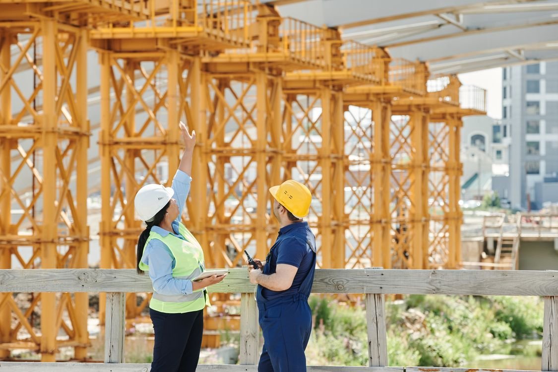 A female engineer in a hard hat and hi-vis vest and a male site worker in a hard hat discussing over a tablet on a construction site, representing the on-site English that construction and site teams rely on