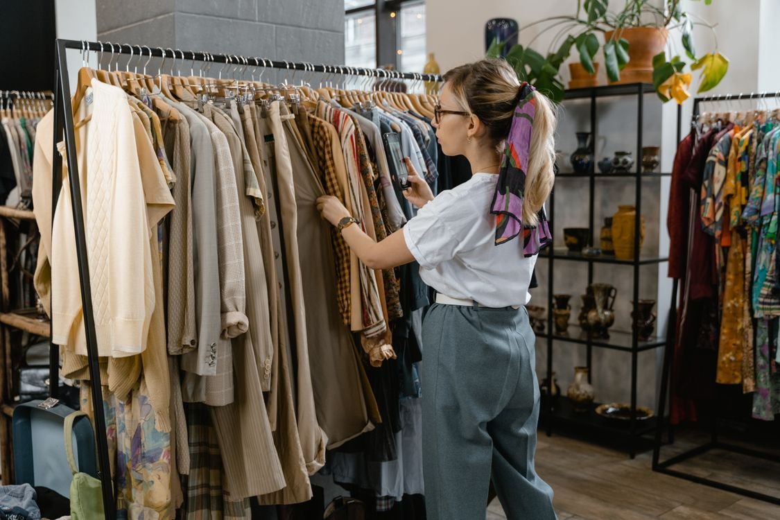 A retail worker on the shop floor of a clothing store looking through a rail of garments while checking her phone, representing the everyday customer, product and stock English shop-floor teams use