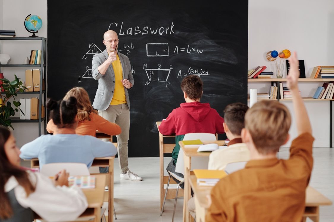 A teacher standing at a blackboard in front of a class of students, representing the everyday classroom English teachers and school staff use