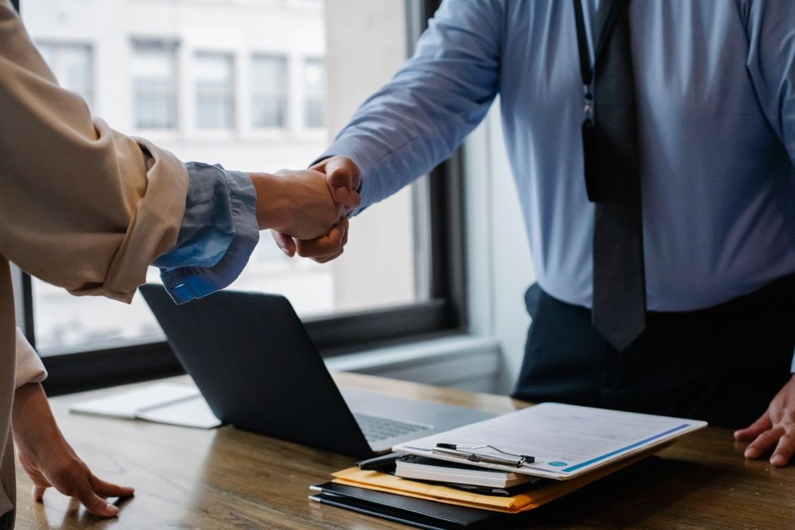 Two professionals shaking hands across a desk at the end of a job interview, with a laptop and interview notes on the table