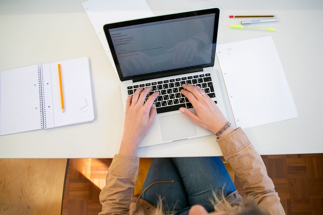 Overhead view of an adult studying English at a laptop on a white desk, with an open spiral notebook and pens beside them