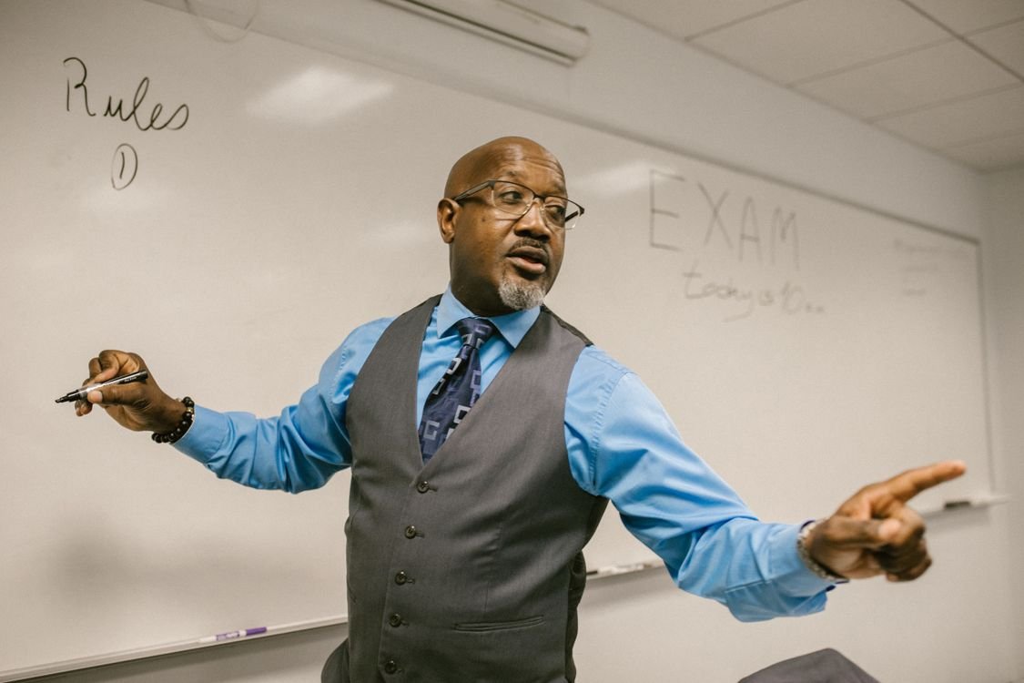 A teacher in a shirt, waistcoat and tie standing at a whiteboard with the word Rules written on it, explaining a grammar point to a class