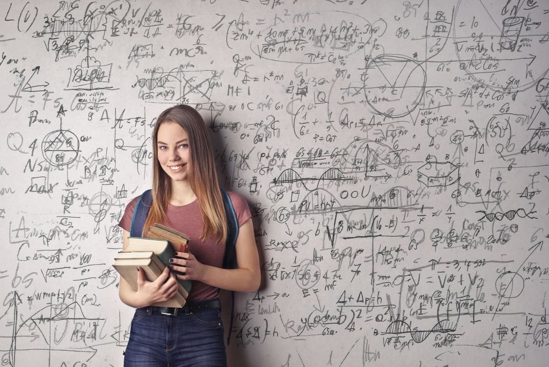 A smiling student holding a stack of books in front of a wall covered in handwritten notes