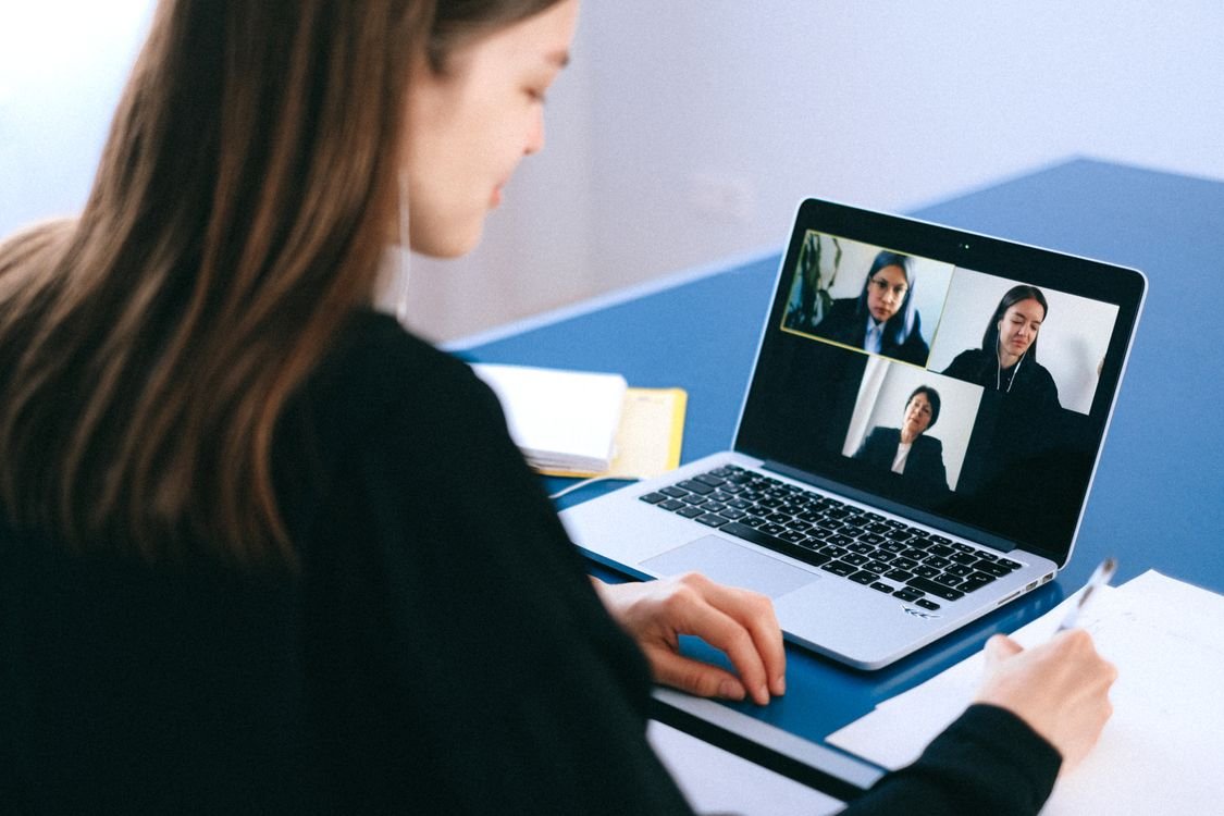 A job candidate at a laptop during a video interview, taking notes while three interviewers appear on the screen