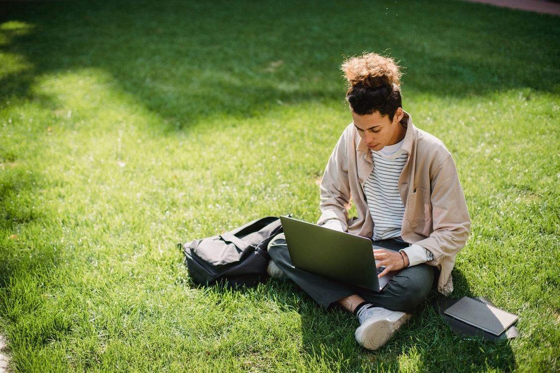 Un adulto joven estudia ingl&eacute;s en l&iacute;nea con el port&aacute;til y un cuaderno, sentado al aire libre en un parque