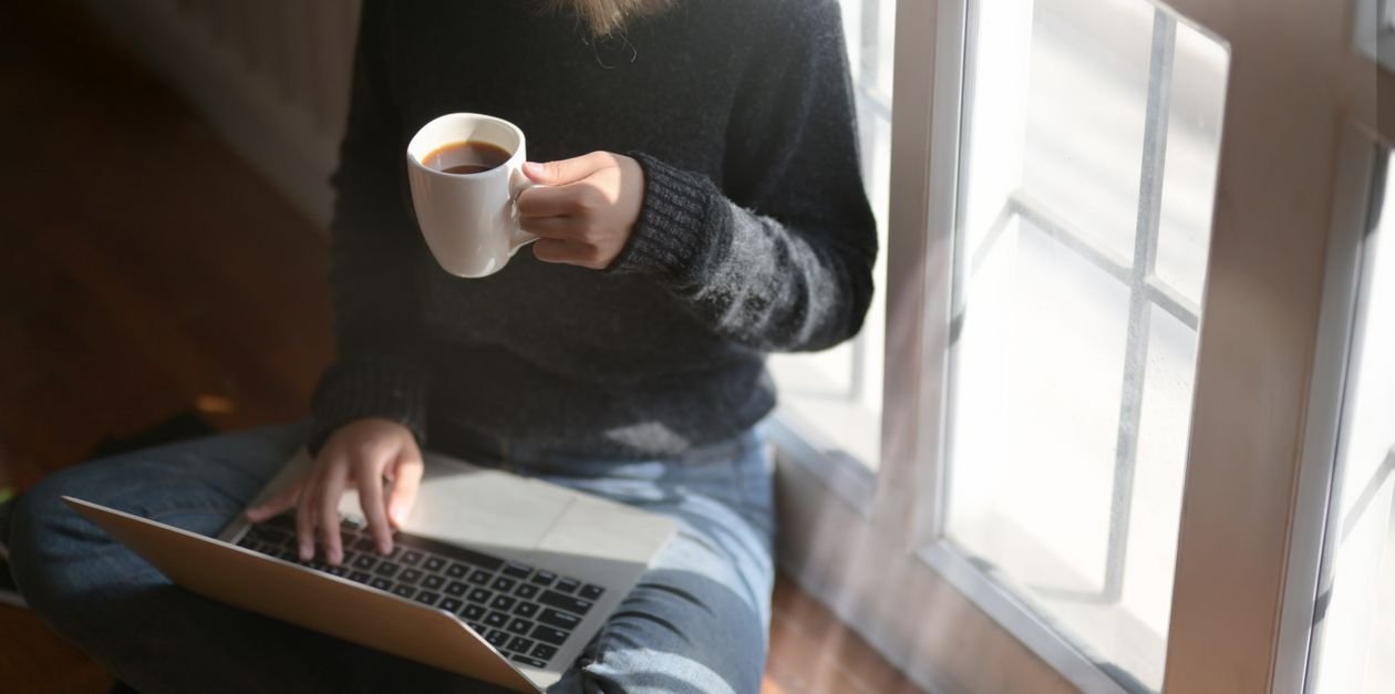 A working adult studying English on a laptop with a cup of coffee by a window at home