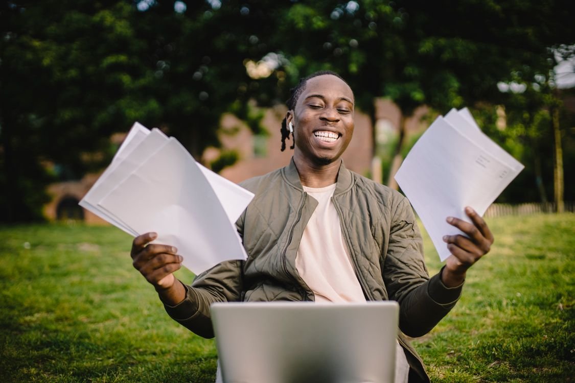 A smiling young adult sitting outdoors with a laptop, holding exam result papers after improving their IELTS score
