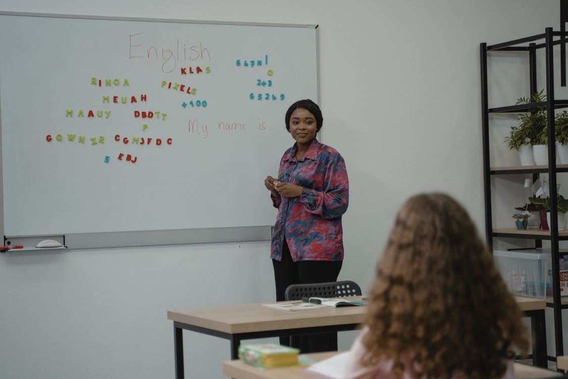 An English teacher standing beside a whiteboard that reads English while an adult learner sits at a desk in a language classroom