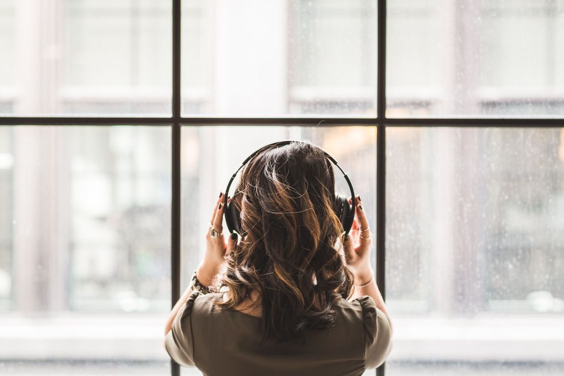 A woman wearing over-ear headphones, listening carefully as she looks out of a window