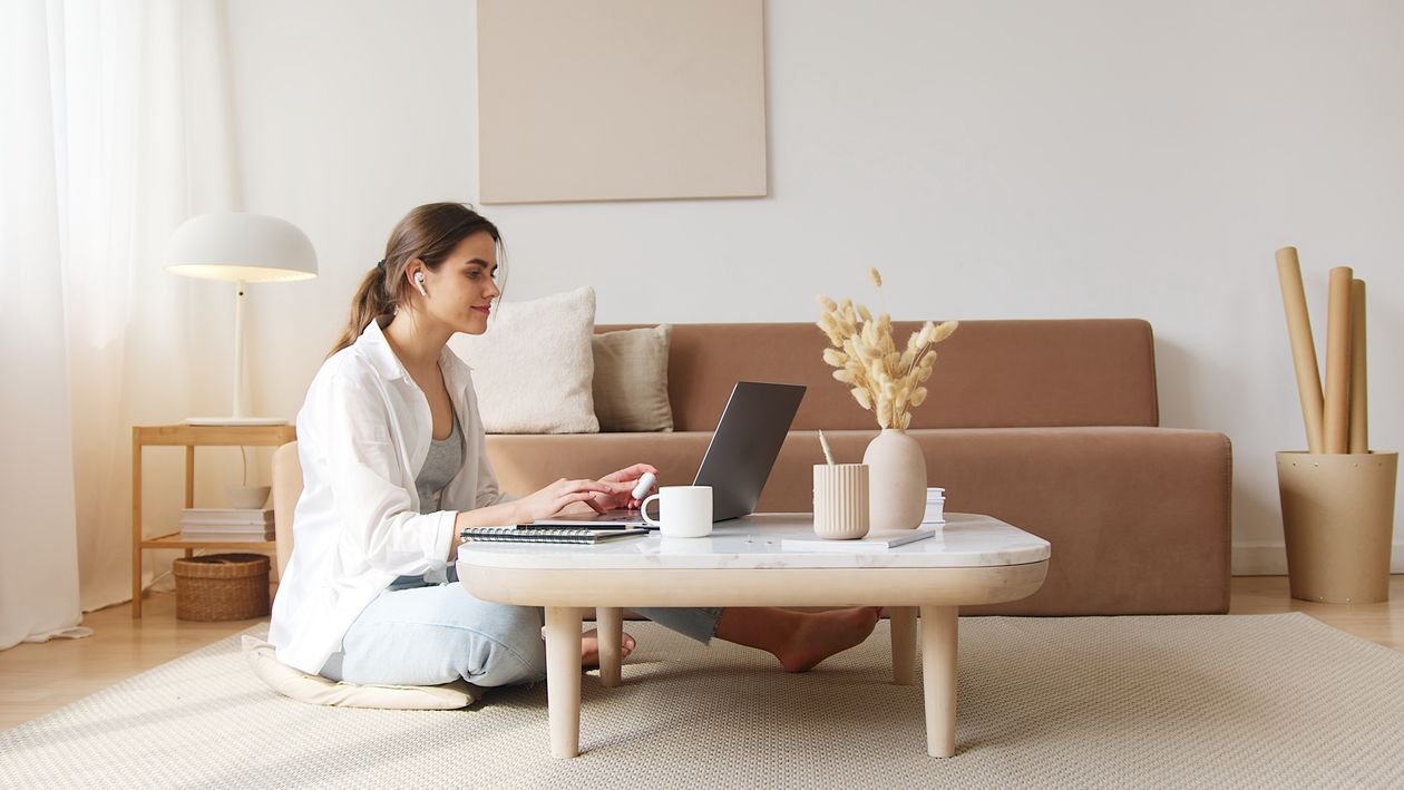 Una donna adulta segue un corso di inglese online da casa, con il portatile e le cuffie, accanto a un quaderno e una tazza di caffè