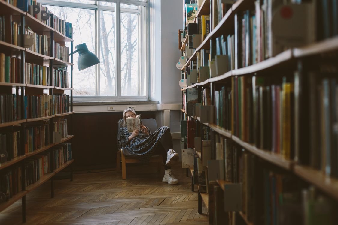 A learner reading a book in a quiet library aisle lined with shelves, absorbed in the story