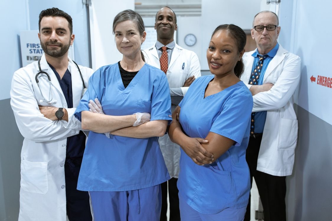 A diverse team of doctors in white coats with stethoscopes and nurses in blue scrubs standing together in a hospital corridor