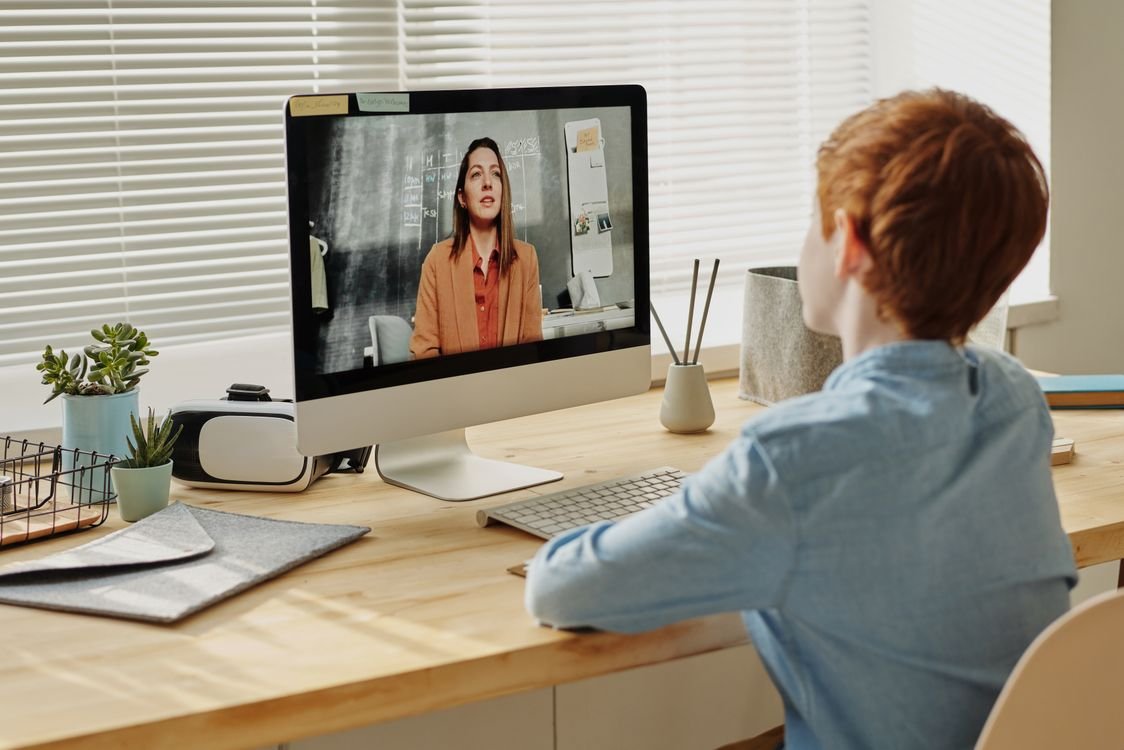 A student in a private one-to-one online English lesson, taking notes at a laptop during a video call with a tutor