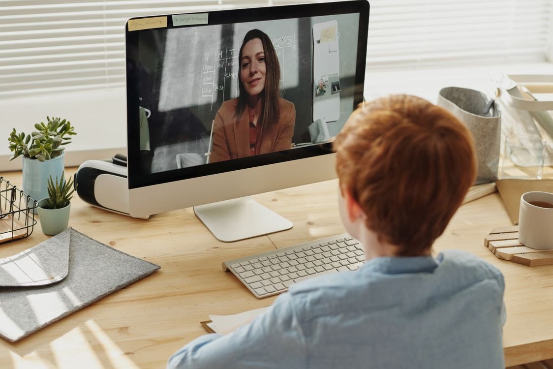 A young student in a live online English lesson, on a video call with a teacher shown on the computer screen at a home desk