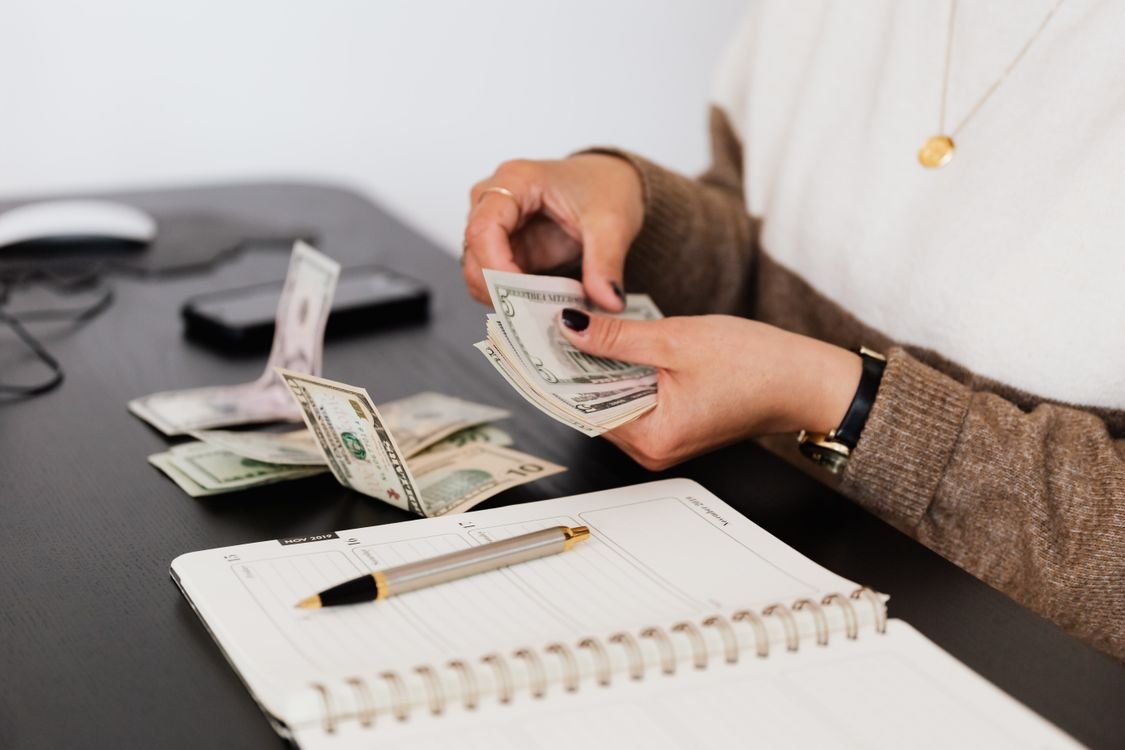 Someone counting banknotes at a desk beside an open budgeting notebook and pen