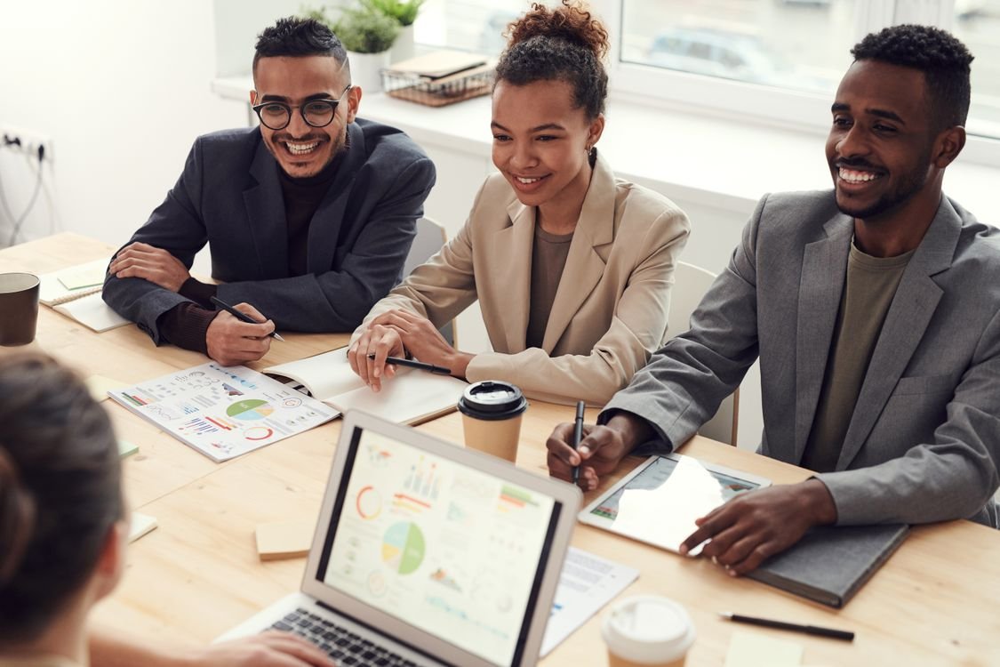 Three professionals in business attire smiling around a table in a workplace meeting, with a laptop showing charts and printed reports